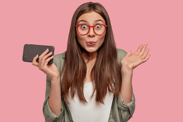 Fototapeta premium Horizontal portrait of attractive brunette girlfriend looks with amazed cheerful expression, holds modern gadget, gossips with close friend about boys, wears round spectacles, oversized shirt