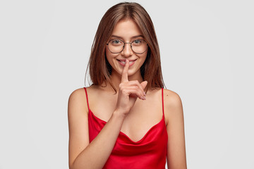 Horizontal shot of secret girl shows silence gesture, keeps index finger on lips, asks not spread rumors, awakes in morning, wears casual nightwear, poses against white background. Silence concept