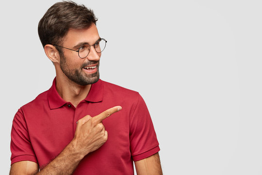 Try that direction. Cheerful unshaven guy wears spectacles, shows way, points aside, has happy satisfied expression, wears red t shirt, isolated over white background, demonstrates free space