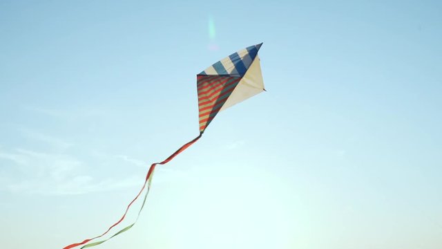 A bright kite with a long tail of silk ribbons flies in a light wind against the blue sky in the rays of the sun at sunset of the day. The Symbol of Liberty and Childhood