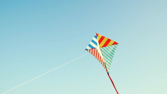 A bright kite with a long tail of silk ribbons flies in a light wind against the blue sky in the rays of the sun at sunset of the day. The Symbol of Liberty and Childhood