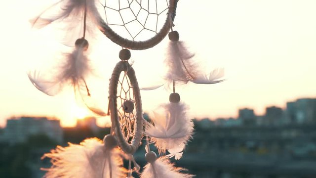 Slow motion amulet dreams catcher waving in a light wind against the backdrop of a bright sunset and a bokeh of city lights. White light feathers rise in the wind. The disc of the setting sun. Ethnic