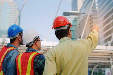 back view group of engineer, technician and architect with safety helmet planning about building plan with blueprint in modern city building background, construction site, industry and worker concept