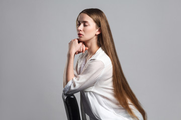 Fototapeta premium side profile view of calm beautiful young model actor with long brown hair in white t-shirt sitting with closed eyes and touching her chin with serious face. studio shot isolated on white background.