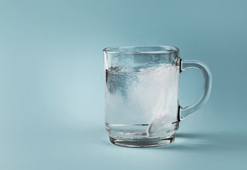 Side view of soluble pain killer dissolving bubbling in water, whole clear glass cup, isolated on blue background. Copy space.
