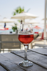 Glass of rose wine with blurred background. Summer wine day view of half-full glass of alcoholic drink on a wooden table with blurred beach restaurant view background and copy space.