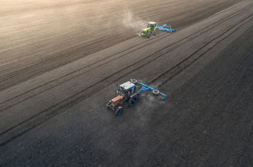 Aerial view of two blue tractors plows the earth in field on a summer day against a black earth...