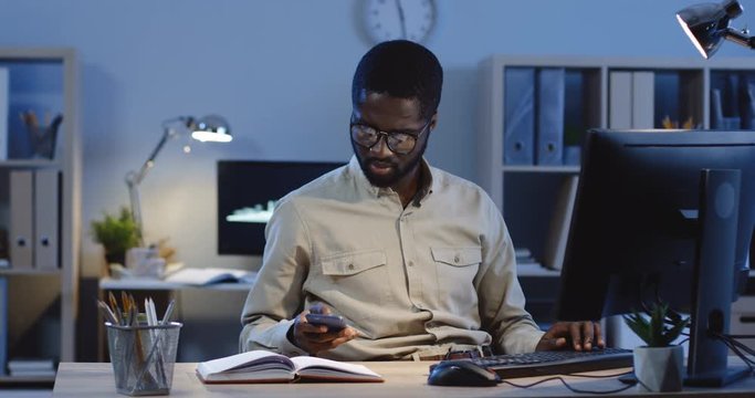 Young attractive African American man working on the computer in the office late at night when getting a message on his smartphone and it cheering him up.