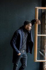 Young black man in coat on dark background. Professional photo studio. Fashion portrait of young man in black shirt in studio. Looking out the window.