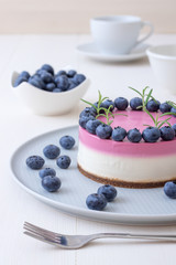 The two-color blueberry mini cheese cake. Round no bake cheesecake, bowl with blueberries, cup of coffee and bowl with sugar. Top of cake decorated by rosemary herbs. White table.