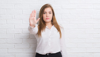 Young adult business woman over white brick wall with open hand doing stop sign with serious and confident expression, defense gesture