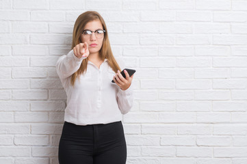 Young adult business woman over white brick wall sending message using smartphone pointing with finger to the camera and to you, hand sign, positive and confident gesture from the front