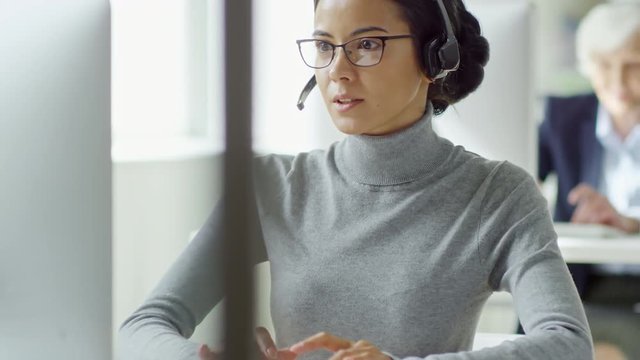 Medium shot of attractive young woman in glasses and headset having conversation with customer while working in call center
