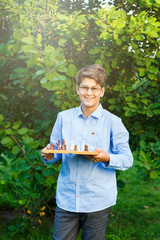 cute young boy in blue shirt holds chess board  and plays chess in the summer park. Education, intellectual game 