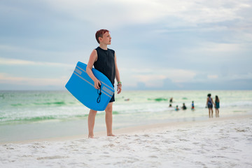 Young boy with wave board on white sand beach looking to future