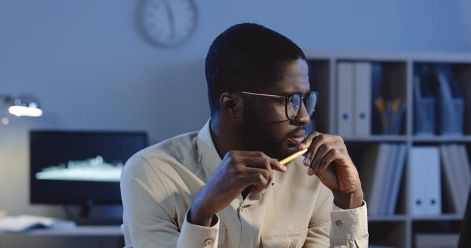 Portrait of the serious African American man in glasses looking at the side, thinking and spinning a pencil in a hand. Late in the office.