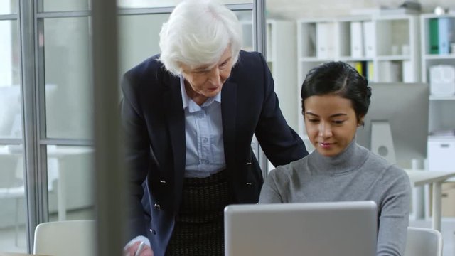 Tilt down shot of two diverse office workers discussing project in office, mature woman helping younger colleague, who is working with laptop computer