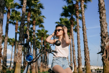 Obraz premium Cheerful female in trendy summer clothes and sunglasses sitting on electrical bicycle over city tropical palm garden background in a sunny day.