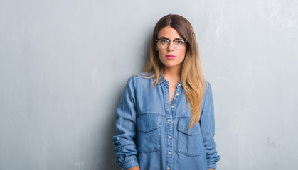 Young adult woman over grunge grey wall wearing glasses with serious expression on face. Simple and...