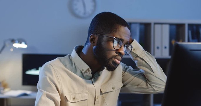 Close up of the young African American office worker in glasses sitting in the office in front of the computer late in the evening, being mixed-up and sad about his work.