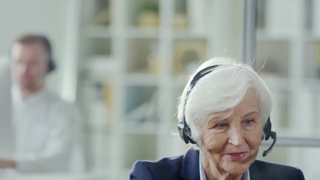 Medium shot of confident senior woman in headset using computer and helping client while working in customer service