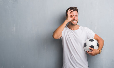 Handsome young man over grey grunge wall holding soccer football ball with happy face smiling doing...