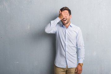 Handsome young business man over grey grunge wall wearing elegant shirt smiling and laughing with hand on face covering eyes for surprise. Blind concept.