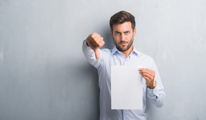 Handsome young man over grey grunge wall holding blank paper sheet contract with angry face,...