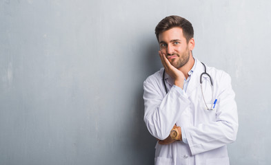 Handsome young doctor man over grey grunge wall thinking looking tired and bored with depression problems with crossed arms.
