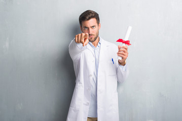 Handsome young doctor man over grey grunge wall holding diploma pointing with finger to the camera and to you, hand sign, positive and confident gesture from the front