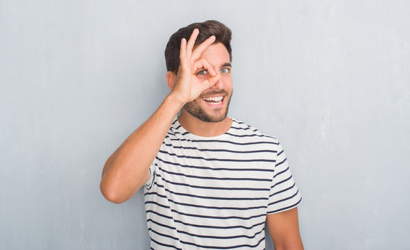 Handsome young man over grey grunge wall wearing navy t-shirt doing ok gesture with hand smiling, eye looking through fingers with happy face.