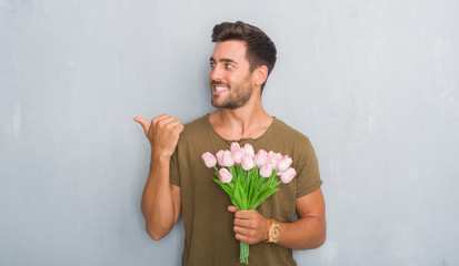 Handsome young man over grey grunge wall holding flowers bouquet pointing and showing with thumb up to the side with happy face smiling