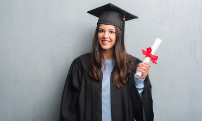 Young brunette woman over grunge grey wall wearing graduate uniform holding degree with a happy...