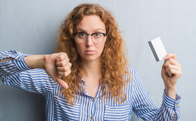 Young redhead woman over grey grunge wall holding credit card with angry face, negative sign showing dislike with thumbs down, rejection concept