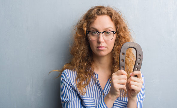 Young redhead woman over grey grunge wall holding horseshoe with a confident expression on smart face thinking serious