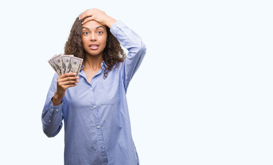 Young hispanic woman holding dollars stressed with hand on head, shocked with shame and surprise face, angry and frustrated. Fear and upset for mistake.