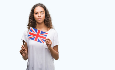 Fototapeta premium Young hispanic woman holding flag of United Kingdom with a confident expression on smart face thinking serious