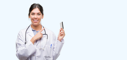 Young hispanic doctor woman holding credit card very happy pointing with hand and finger to the side