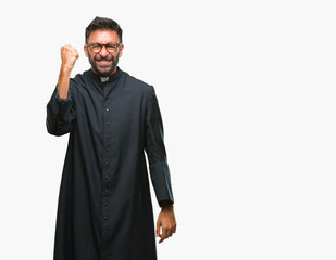Adult hispanic catholic priest man over isolated background angry and mad raising fist frustrated...