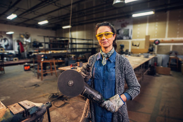 Fototapeta na wymiar Picture of professional middle age female carpenter standing in her workshop and holding electric drill. Looking at camera and smiling.
