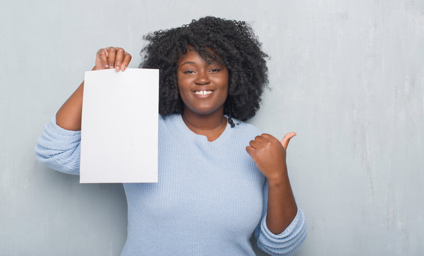 Young african american woman over grey grunge wall holding blank paper sheet pointing and showing with thumb up to the side with happy face smiling
