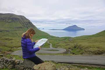 Naklejka premium CLOSE UP: Young female tourist reading a map while sitting on a rock by the road