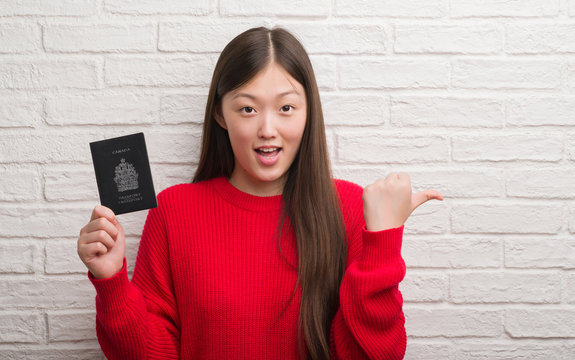Young Chinese woman over brick wall holding passport of Canada pointing and showing with thumb up to the side with happy face smiling