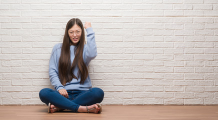 Young Chinese woman sitting on the floor over brick wall angry and mad raising fist frustrated and furious while shouting with anger. Rage and aggressive concept.