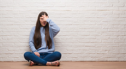 Young Chinese woman sitting on the floor over brick wall peeking in shock covering face and eyes with hand, looking through fingers with embarrassed expression.