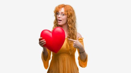 Young redhead woman in love holding red heart very happy pointing with hand and finger