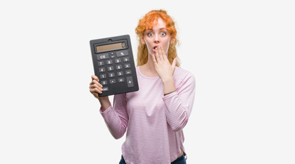 Young redhead woman holding big calculator cover mouth with hand shocked with shame for mistake,...
