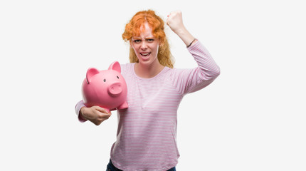 Young redhead woman holding piggy bank annoyed and frustrated shouting with anger, crazy and yelling with raised hand, anger concept
