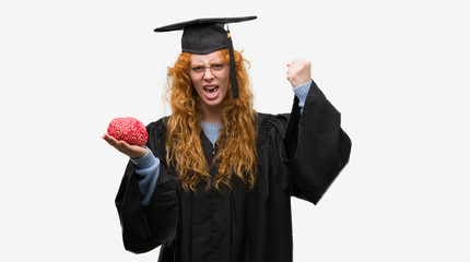 Young redhead student woman wearing graduated uniform holding brain annoyed and frustrated shouting with anger, crazy and yelling with raised hand, anger concept