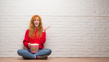 Young redhead woman sitting over brick wall eating popcorn very happy and excited, winner expression celebrating victory screaming with big smile and raised hands